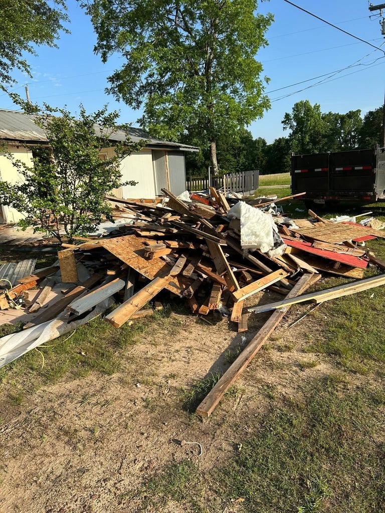 Pile of scrap wood and metal debris scattered in a rural yard with buildings and trees in the background