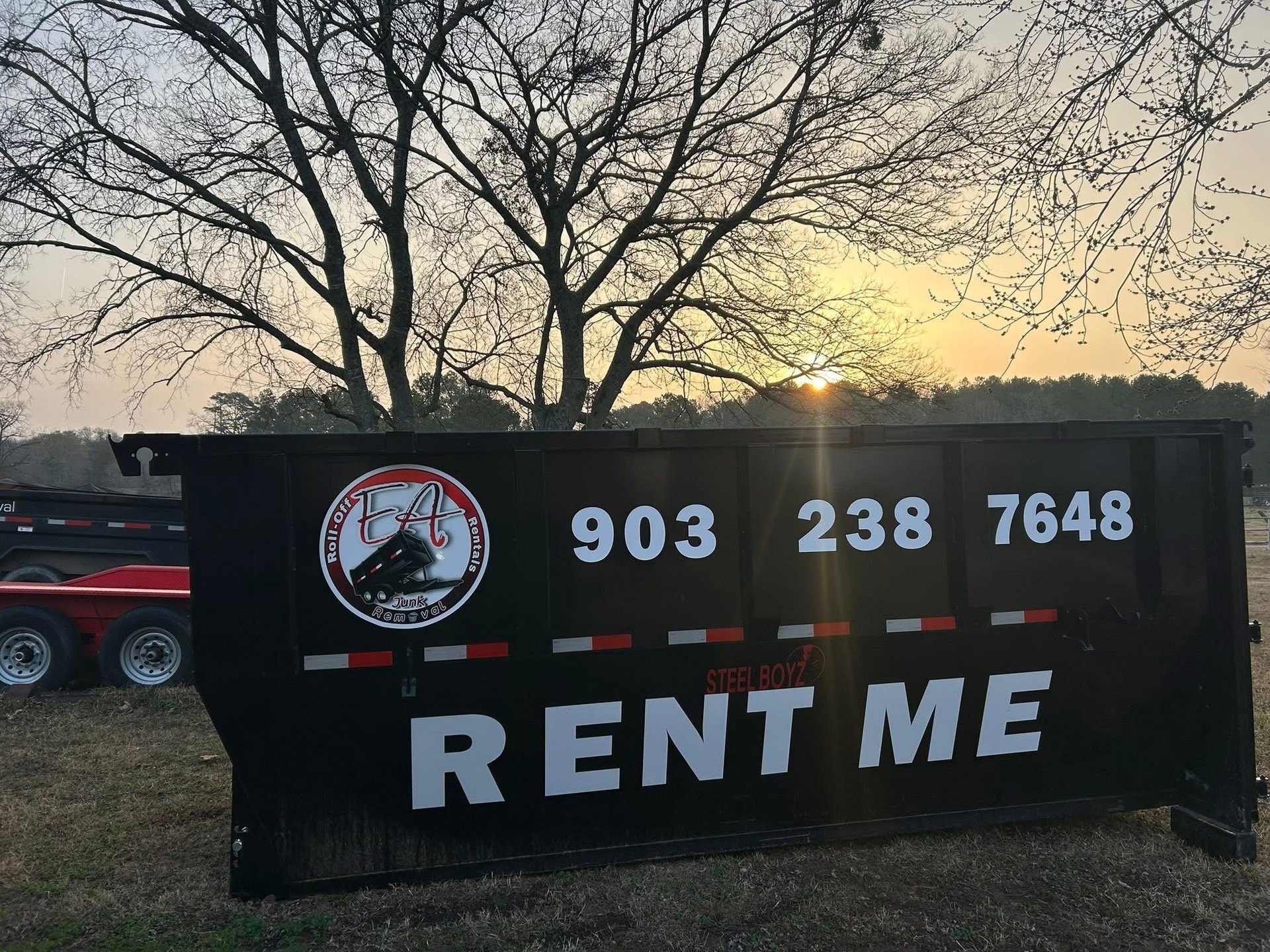 Black rental dumpster trailer with phone number and RENT ME text, parked outdoors at sunset with bare trees in background