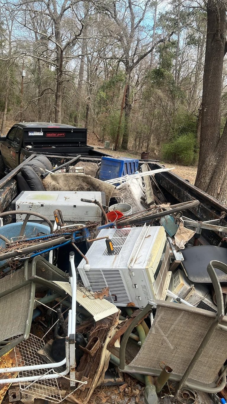 A pickup truck bed filled with discarded appliances and junk in a wooded area, including an air conditioning unit and other debris
